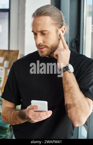 uomo d'affari premuroso con cuffie regolabili da smartphone mentre ascolta il podcast in ufficio Foto Stock