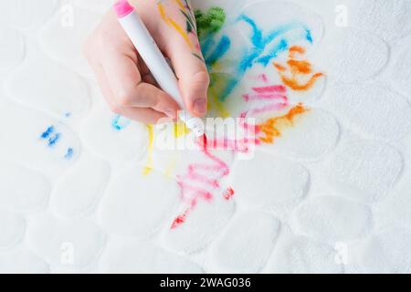 La mano di un bambino disegna penne in feltro sui mobili imbottiti bianchi del divano o di un tappeto. vista dall'alto. Macchia sporca per il concetto di pulizia. Foto Stock