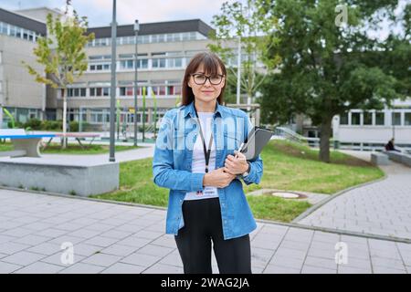 Donna di mezza età insegnante di scuola con un tablet digitale che posa vicino all'edificio scolastico Foto Stock