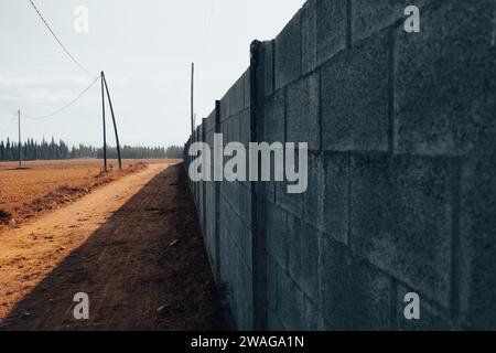 Strada polverosa di terra rossa accanto a un muro di mattoni, con cipressi all'orizzonte sotto un cielo biancastro, che suggerisce un'atmosfera distopica Foto Stock