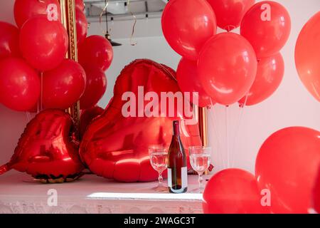 Sul tavolo bianco bottiglia di vino con due bicchieri tra palloncini rossi gonfiabili a forma di cuore. Biglietto d'auguri creativo per San Valentino, compleanno, Foto Stock