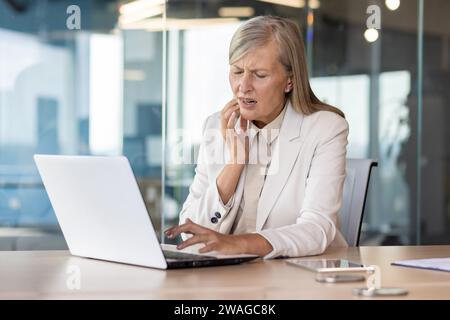 Un'anziana donna d'affari dai capelli grigi è seduta a una scrivania in ufficio, lavora su un notebook, tiene la mano sulla guancia, soffre di un forte mal di denti. Foto Stock