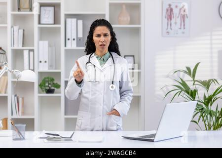 Giovane medico latinoamericana in piedi in uno studio clinico vicino alla scrivania, guardando seriamente la macchina fotografica e mostrando il dito che proibisce gesti. Foto Stock