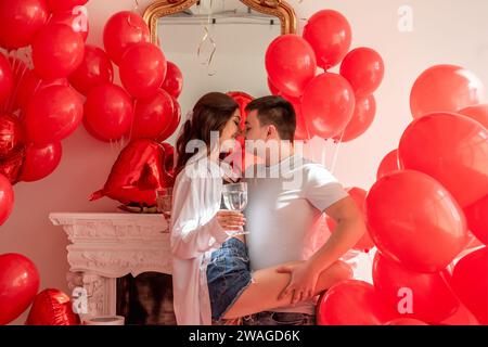 Un momento di gioia tra la danza di giovani coppie che festeggiano con un brindisi di San Valentino vicino a palloncini rossi. Donna che regge un bicchiere di vino, occhi coperti, uomo abbraccia, Foto Stock