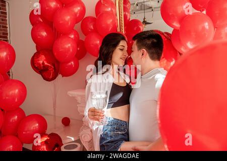 Un momento di gioia tra la danza di giovani coppie che festeggiano con un brindisi di San Valentino vicino a palloncini rossi. Donna che regge un bicchiere di vino, occhi coperti, uomo abbraccia, Foto Stock