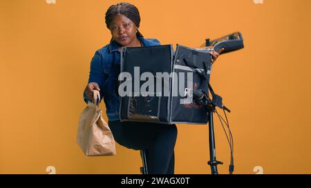 Donna afro-americana che afferra il cibo da asporto dal sacchetto per la consegna a domicilio per persona nel quartiere locale. Corriere sorridente che offre un servizio clienti rapido e affidabile, offrendo al cliente pasti da asporto freschi. Foto Stock