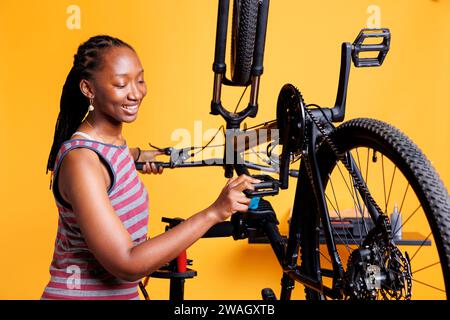 Giovane ciclista attiva assicura che la bici sia ben preparata per le avventure all'aria aperta esaminando diligentemente i pedali e le attrezzature. Donna afroamericana sportiva che afferra la leva a pedale della bicicletta per la manutenzione. Foto Stock