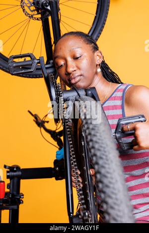 Donna nera dedicata che analizza da vicino il soggiorno della catena di bici per mantenere l'efficienza del ciclo. Ciclista afroamericana che si occupa della riparazione di catene portarulli per biciclette con attrezzature specializzate. Foto Stock