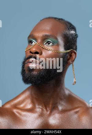 Uomo gay barbato afro-americano sicuro di sé con un trucco luminoso isolato su sfondo blu, ritratto ravvicinato. Trasuda senso di orgoglio e individualità. Potere di diversità dello stile personale. Foto Stock