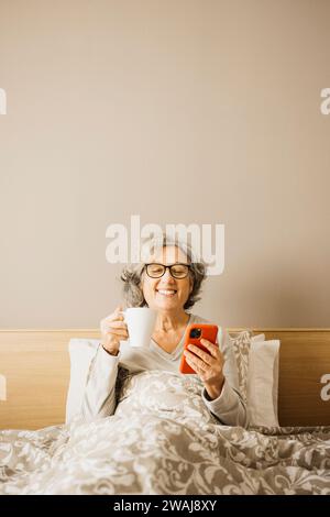 Donna anziana seduta a letto la mattina con pigiama e bevendo caffè con una tazza bianca mentre controlla il telefono. Foto Stock
