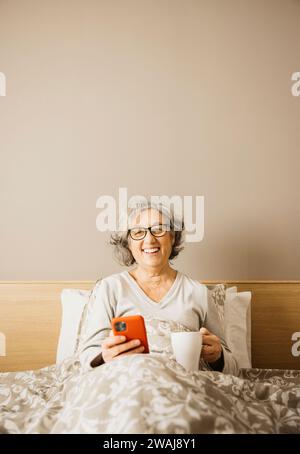 Ritratto di una donna anziana seduta a letto la mattina con pigiama e caffè bevuto con una tazza bianca mentre controlla il telefono. Foto Stock