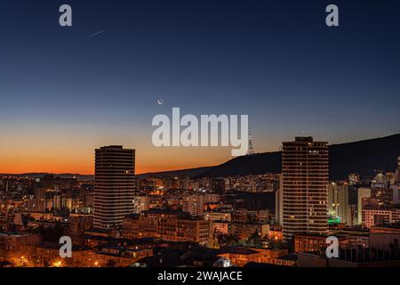 La luna a mezzaluna pende nel cielo crepuscolo mentre l'alba si apre sullo skyline del centro di Tbilisi illuminando la città in un caldo bagliore Foto Stock
