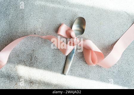 Un cucchiaio in argento legato a un delicato nastro rosa è adagiato su un panno grigio testurizzato, che presenta un dettaglio semplice ma elegante Foto Stock