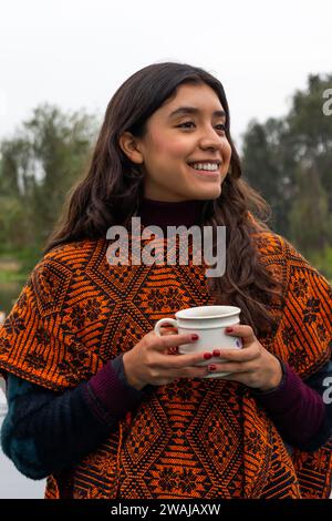 Ritratto donna gioiosa avvolto in un tradizionale scialle decorato regge una tazza sorridente all'aperto con uno sfondo morbido Foto Stock