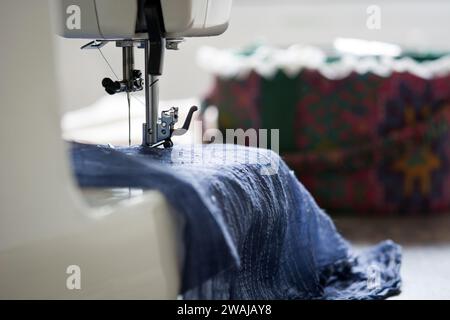 Foto dettagliata di un ago per macchina da cucire e del piede presser su denim blu, con un cuscino colorato a spillo sullo sfondo. Foto Stock