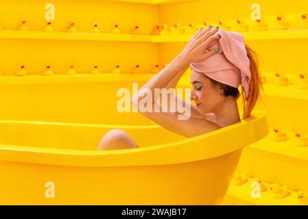 La vista laterale della donna si rilassa con un'espressione contemplativa in una vasca da bagno gialla, un asciugamano avvolto intorno ai suoi capelli sullo sfondo di anatre di gomma Foto Stock