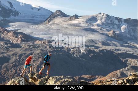 Corpo completo di due corridori che si muovono rapidamente su un sentiero roccioso di montagna con un vasto ghiacciaio e cime innevate sullo sfondo sotto un blu tenue Foto Stock