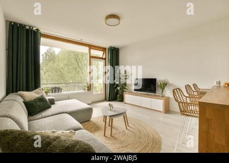 Televisore e divano grigio posti da una grande finestra luminosa in un ampio soggiorno di un appartamento contemporaneo con sedie vicino all'isola Foto Stock