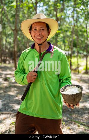 Ritratto dei sorridenti agricoltori asiatici di gomma in piedi con tazze di lattice in una piantagione di alberi di gomma. Foto Stock