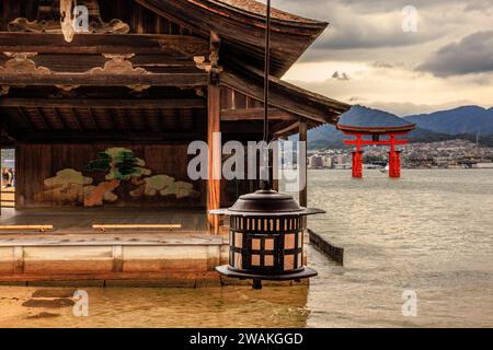 una lanterna di metallo è appesa davanti all'edificio del santuario di itsukushima con la famosa porta torii rossa galleggiante dell'isola di miyajima nella baia di hiroshima sullo sfondo Foto Stock