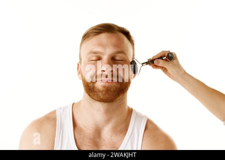 Ritratto ravvicinato di un uomo che ha un messaggio facciale con rullo nel centro termale e che si diverte sullo sfondo bianco dello studio. Procedure di benessere nel centro benessere. Foto Stock