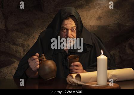 Ritratto emotivo di un monaco in cassock, calice di argilla in mano, seduto a un tavolo, una brocca di vino e bevuto in una locanda medievale. Concetto: Ubriachezza a Foto Stock