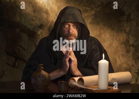 ritratto di un monaco anziano in una veste nera, mani piegate in preghiera, una cella scura, una candela che brucia su un tavolo. Foto Stock