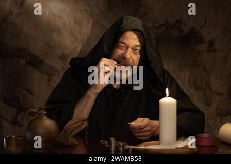 Un monaco autentica il denaro a lume di candela in una stanza di pietra scura, pile di monete. Concetto: Esattore delle tasse nel Medioevo, tesoriere della chiesa. Foto Stock