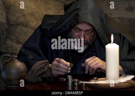 Ritratto di un monaco in una cassoca, contando i soldi a lume di candela in una stanza di pietra scura, pile di monete. Nel Medioevo - esattore delle tasse, chiesa Treas Foto Stock