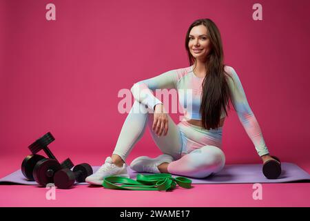 Donna allegra e sportiva che sorride alla fotocamera mentre è seduta sul pavimento in studio. Vista frontale di una donna caucasica di mezza età in abbigliamento sportivo, seduta sul pavimento, circondata da attrezzature sportive. Concetto sportivo. Foto Stock