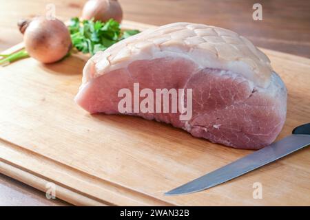 Carne di maiale arrosto salata cruda su un tagliere di legno con coltello da cucina, cipolle ed erbe, preparazione per un prosciutto natalizio con crosta, copia Foto Stock