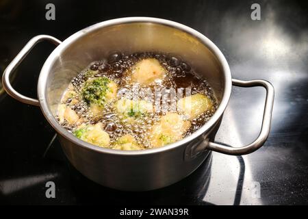 I cavolfiori e i broccoli in pastella vengono fritti in profondità in una pentola con olio bollente caldo su un piano cottura nero, cucinando un gustoso spuntino vegano con verdure, Foto Stock