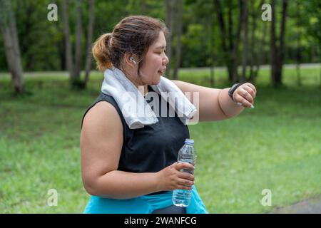 Giovane donna in sovrappeso che controlla il risultato della sua corsa mattutina sul suo orologio intelligente mentre si trova sulla pista da corsa di un parco locale Foto Stock