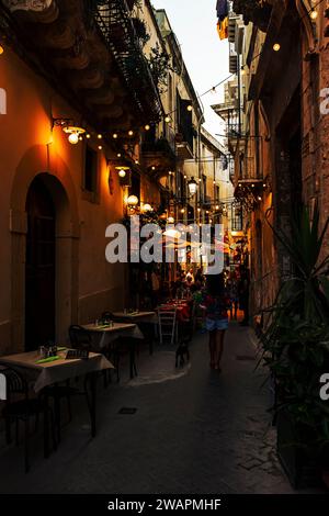 Una donna mediterranea dai capelli ricci cammina al tramonto con il suo cane attraverso i vicoli di Ortigia alla ricerca di un ristorante Foto Stock