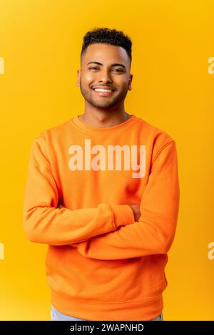 Ritratto di un simpatico e sorridente indiano in posa casual con le braccia incrociate su sfondo arancione, uno studente allegro guarda la fotocamera con un sorriso amichevole Foto Stock