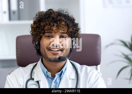 Vista webcam, giovane medico con telefono auricolare utilizzando computer portatile per videochiamata, medico allegro e amichevole consultazione pazienti, sorridente e guardando la macchina fotografica, lavoro all'interno della clinica sala medica. Foto Stock