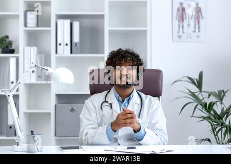 Vista webcam, giovane medico con telefono auricolare utilizzando computer portatile per videochiamata, medico allegro e amichevole consultazione pazienti, sorridente e guardando la macchina fotografica, lavoro all'interno della clinica sala medica. Foto Stock