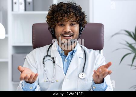 Vista webcam, giovane medico con telefono auricolare utilizzando computer portatile per videochiamata, medico allegro e amichevole consultazione pazienti, sorridente e guardando la macchina fotografica, lavoro all'interno della clinica sala medica. Foto Stock