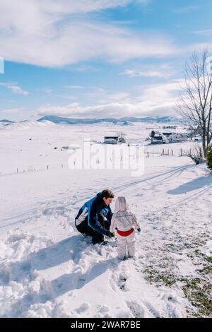 Un bambino piccolo si trova accanto a suo padre, che sta accovacciando nella neve, e lo guarda. Vista posteriore Foto Stock