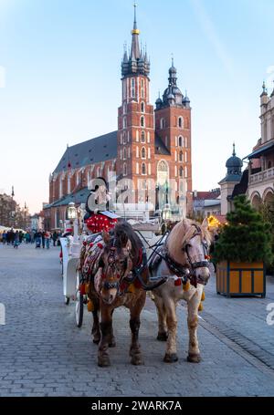 CRACOVIA, POLONIA - 27 DICEMBRE 2023: Carrozza trainata da cavalli durante il periodo natalizio. La piazza principale è circondata da storiche case cittadine (kamienice) e. Foto Stock