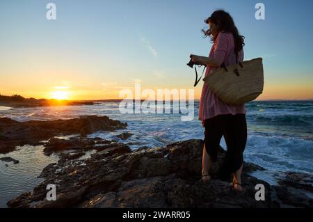 Donna che fotografa al tramonto sulla costa nella spiaggia di Ses Platgetes a es Caló (Formentera, Isole Baleari, Mar Mediterraneo, Spagna) Foto Stock