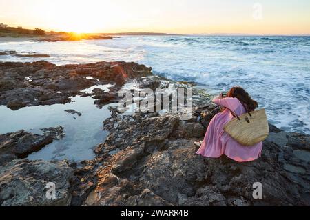 Donna che fotografa al tramonto sulla costa nella spiaggia di Ses Platgetes a es Caló (Formentera, Isole Baleari, Mar Mediterraneo, Spagna) Foto Stock