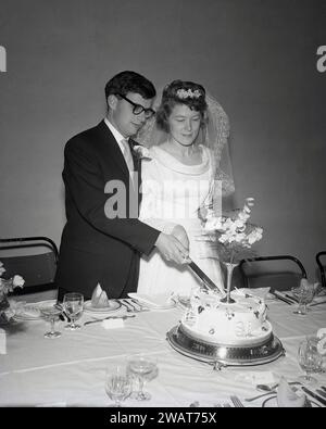 Anni '1950, storico, al ricevimento di nozze, una sposa e uno sposo tagliano insieme la torta di nozze, Inghilterra, Regno Unito. Foto Stock