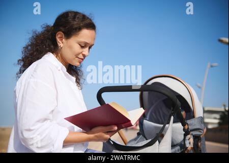 Felice giovane donna latinoamericana, madre che spinge il passeggino, legge un libro, si rilassa sul parapetto sulla spiaggia atlantica. Congedo di maternità. Peopl Foto Stock