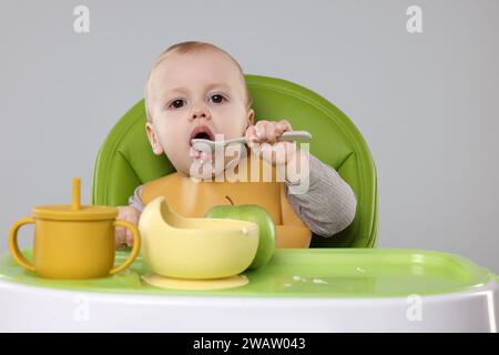 Piccolo bambino carino che mangia cibo sano in seggiolone su sfondo grigio Foto Stock