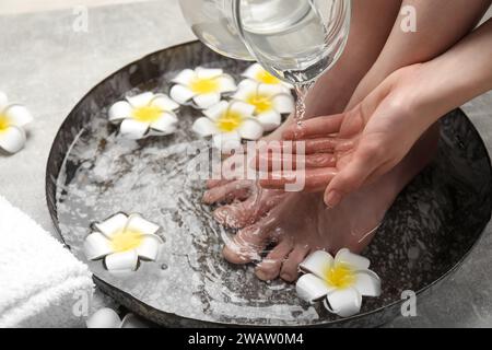 Donna che versa acqua sulla mano mentre ammorbidisce i piedi in un recipiente grigio chiaro, primo piano. Trattamento spa Foto Stock