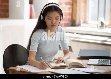 Studente asiatico concentrato che studia prendendo appunti indossando cuffie Foto Stock