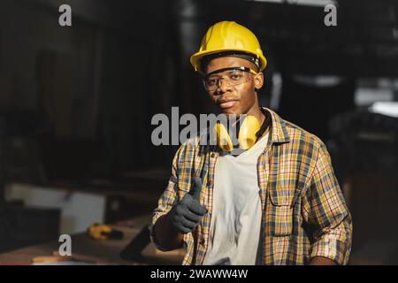 lavoratore nero africano in officina con elmetto di sicurezza e cuffia con pollice in piedi fotocamera Foto Stock