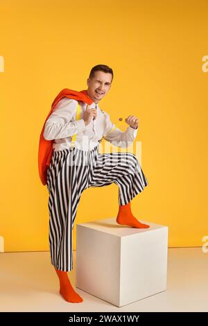 il buffo pazzo con giacca arancione e pantaloni a righe gli regala un paio di bretelle su sfondo giallo Foto Stock