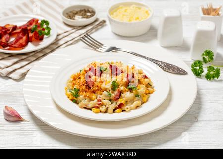 porzione di mac e formaggio con mais, pancetta ricoperta di pangrattato panko su piatto bianco su tavola di legno bianco con ingredienti sullo sfondo, orizzonte Foto Stock
